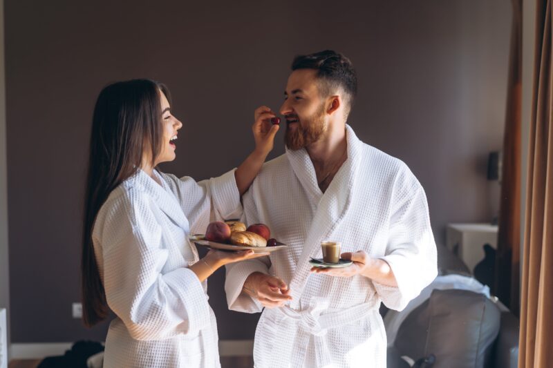 Nasmejani par u bademantilima uživa u doručku uz kafu i voće u opuštenoj atmosferi hotelske sobe.