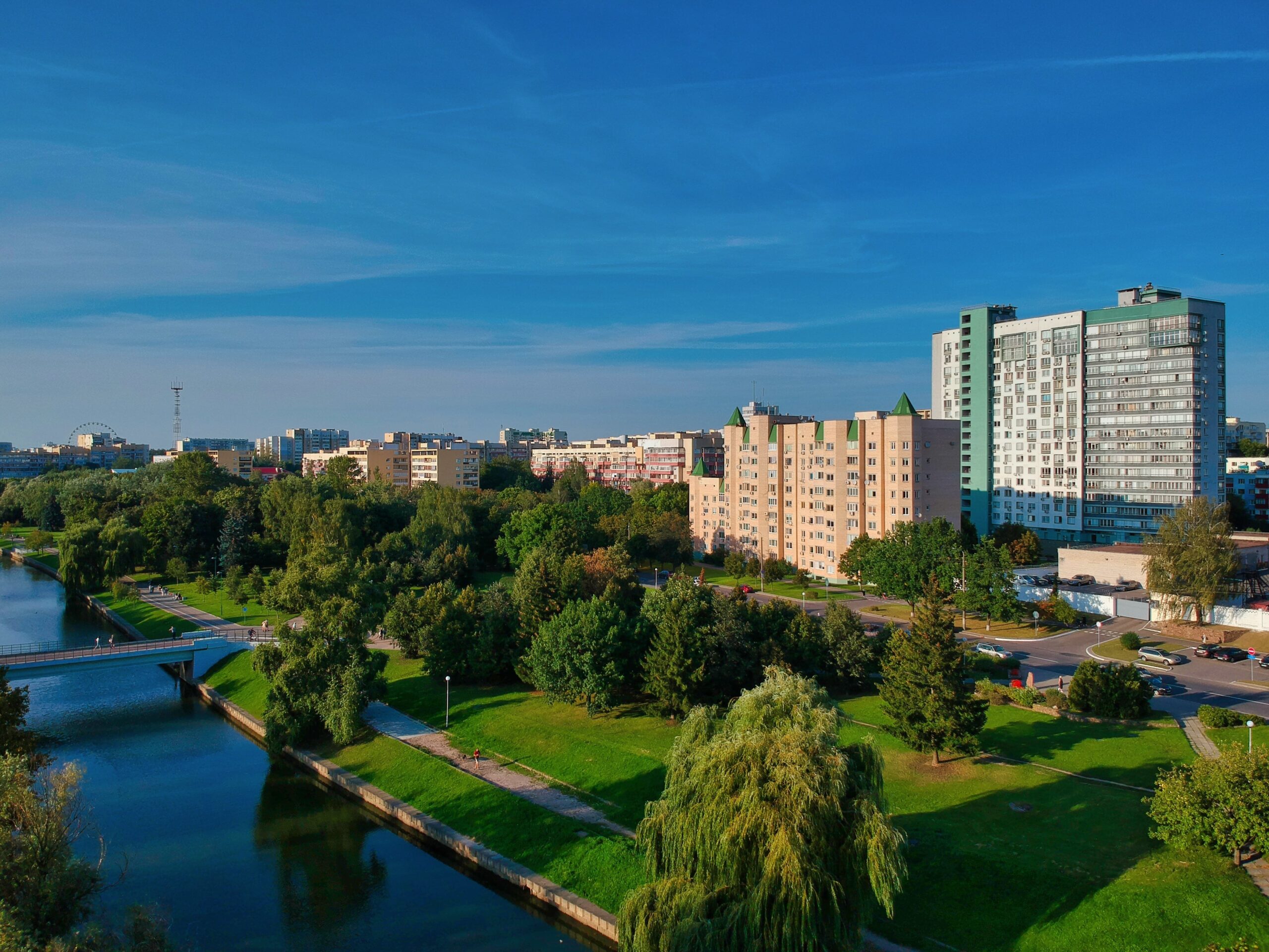 Panoramski pogled na Novi Sad sa modernim stambenim zgradama, zelenim parkovima i rekom, pod vedrim plavim nebom.